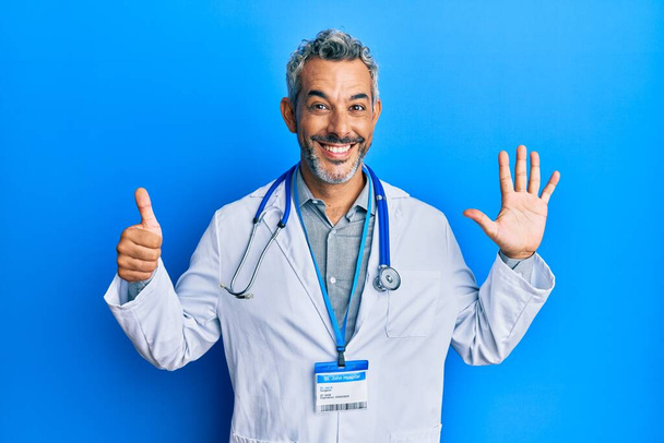 Középkorú ősz hajú férfi orvosi egyenruhában és sztetoszkóppal a hatodik ujjal mutogatva, miközben magabiztosan és boldogan mosolyog..  - Fotó, kép