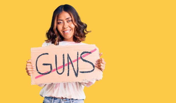 Young beautiful mixed race woman holding no guns warning banner looking positive and happy standing and smiling with a confident smile showing teeth  - Photo, Image