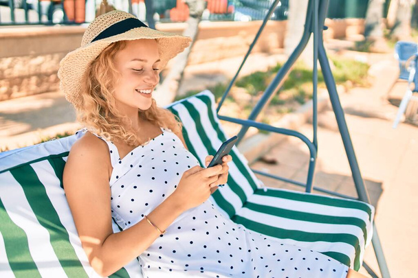 Νεαρή όμορφη καυκάσια γυναίκα με ξανθά μαλλιά χαμογελώντας ευτυχισμένη σε εξωτερικούς χώρους χρησιμοποιώντας το smartphone - Φωτογραφία, εικόνα