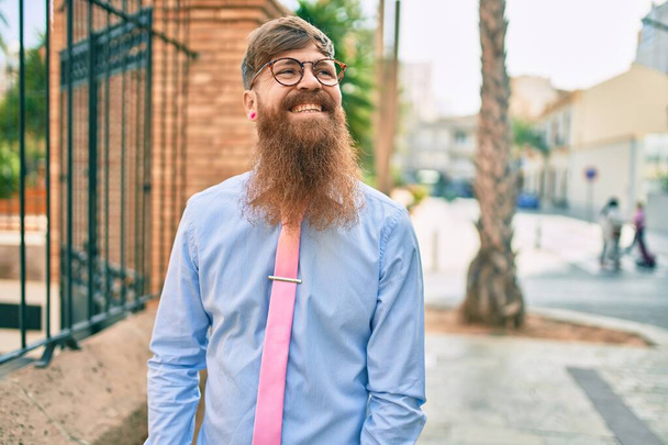 Joven hombre de negocios pelirroja con barba larga sonriendo feliz caminando por la ciudad. - Foto, Imagen