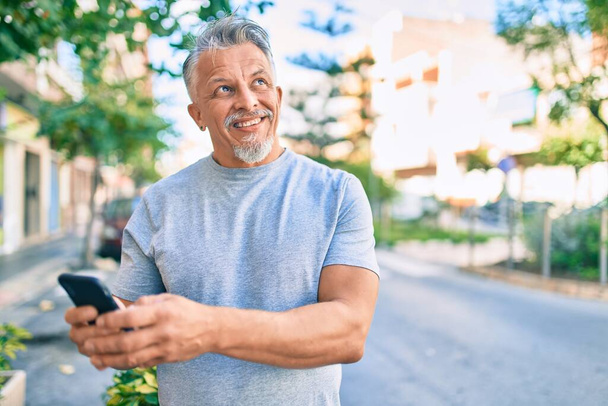 Hombre hispano de pelo gris de mediana edad sonriendo feliz usando el teléfono inteligente en la ciudad. - Foto, imagen