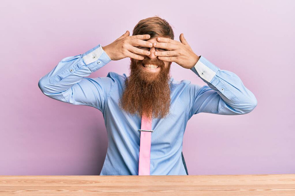 Junger irischer Rotschopf in Businesshemd und Krawatte sitzt auf dem Tisch und bedeckt die Augen mit Händen, die fröhlich und lustig lächeln. Blindes Konzept.  - Foto, Bild