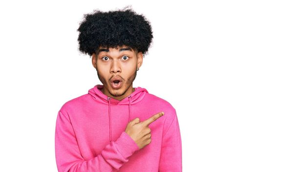 Jeune homme afro-américain aux cheveux afro portant un sweat-shirt rose décontracté surpris pointant du doigt sur le côté, bouche ouverte expression étonnante.  - Photo, image