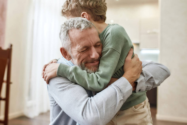 Emotional meeting. Happy grandfather embracing his cute little preschool grandson while standing together at home - Fotoğraf, Görsel