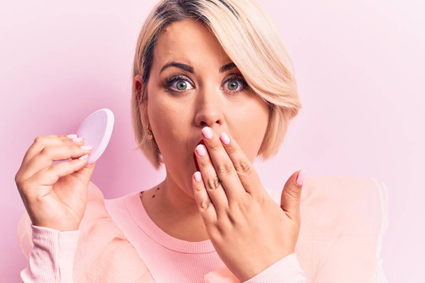 Jovem linda loira plus size mulher aplicando maquiagem usando esponja sobre fundo rosa cobrindo a boca com a mão, chocado e com medo de erro. Expressão surpreendida - Foto, Imagem