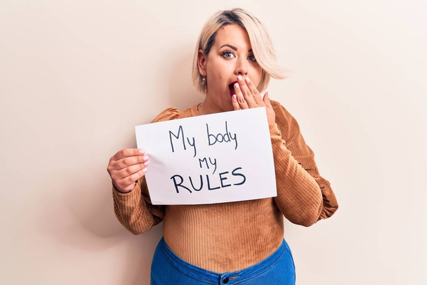 Blonde plus size woman asking for women rights holding paper with my body my rules message covering mouth with hand, shocked and afraid for mistake. Surprised expression - Foto, Imagem