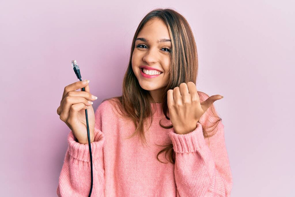 Jeune femme brune tenant un câble Ethernet rj45 pointant le pouce vers le côté souriant heureux avec la bouche ouverte  - Photo, image
