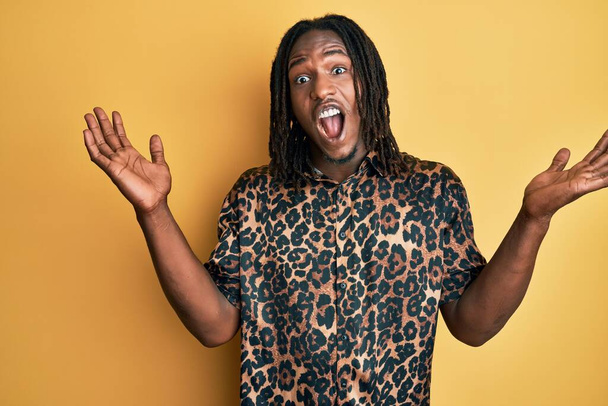 Homme afro-américain avec des tresses portant une chemise léopard imprimé animal célébrant fou et étonné pour le succès avec les bras levés et les yeux ouverts hurlant excités. concept gagnant  - Photo, image