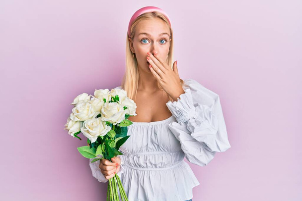 Young blonde girl holding flowers covering mouth with hand, shocked and afraid for mistake. surprised expression  - Photo, Image