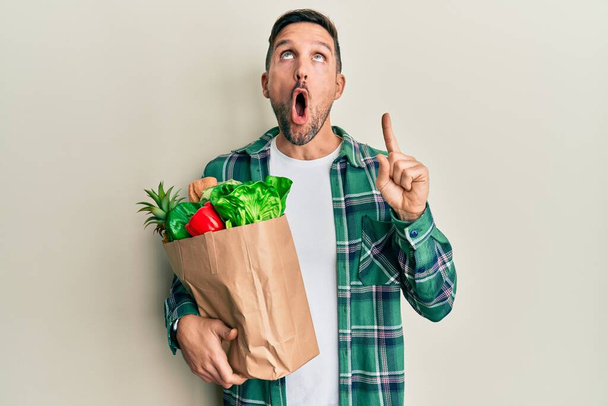 Pohledný muž s vousy drží papírový sáček s potravinami ohromený a překvapený vzhlíží a ukazuje s prsty a zvednutými pažemi.  - Fotografie, Obrázek