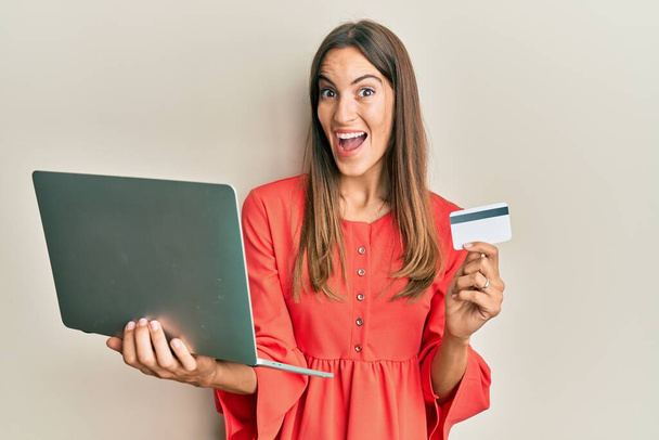 Young beautiful woman holding laptop and credit card celebrating crazy and amazed for success with open eyes screaming excited.  - 写真・画像