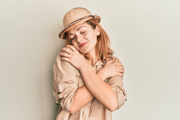 Kafkasyalı genç bir kadın kaşif şapkası takıyor mutlu ve olumlu bir şekilde kendine sarılıyor, kendinden emin gülümsüyor. Sevgi ve özgüven.  - Fotoğraf, Görsel