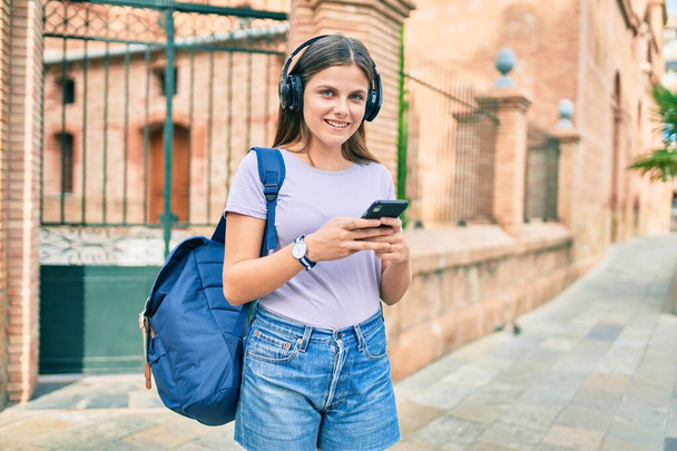 Młoda studentka z Bliskiego Wschodu uśmiechnięta za pomocą słuchawek i smartfona na kampusie uniwersyteckim. - Zdjęcie, obraz