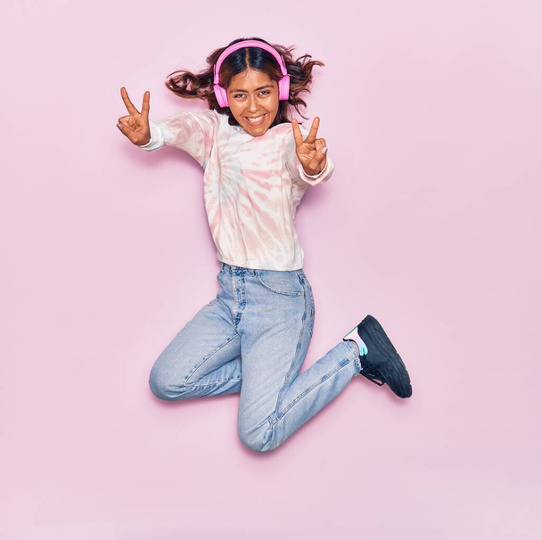 Young beautiful latin woman listening to music using headphones smiling happy. Jumping with smile on face doing victory sign over isolated pink background - Foto, Imagem
