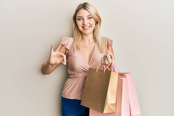 Młoda kaukaska kobieta trzyma torby na zakupy robi ok znak palcami, uśmiechnięty przyjazny gest doskonały symbol  - Zdjęcie, obraz