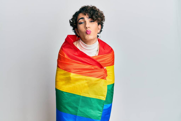 Junger Mann mit Frauenschminke, in Regenbogenflagge gehüllt, blickt in die Kamera und bläst einen Kuss, der schön und sexy ist. Liebesausdruck.  - Foto, Bild