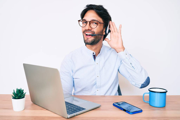 Hombre hispano guapo que trabaja en la oficina con auriculares de operador sonriendo con la mano sobre el oído escuchando y escuchando rumores o chismes. concepto de sordera.  - Foto, imagen