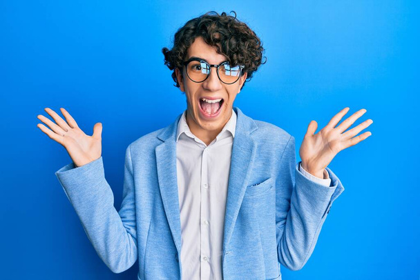Hispánský mladý muž v obchodní bundě a brýlích slaví vítězství se šťastným úsměvem a vítězným výrazem se zdviženýma rukama  - Fotografie, Obrázek