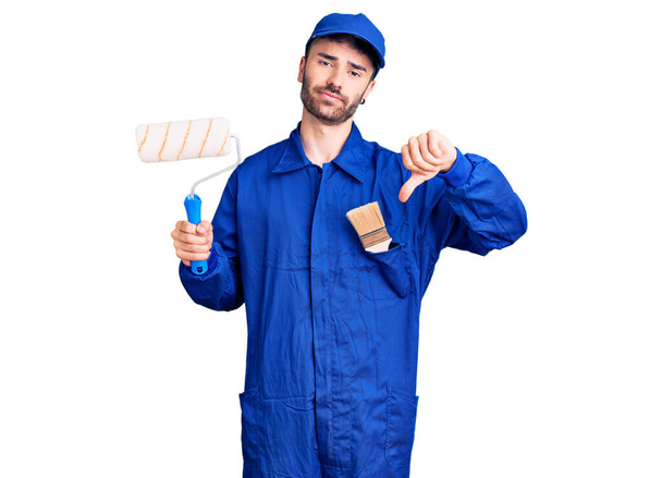 Young hispanic man wearing painter uniform holding roller with angry face, negative sign showing dislike with thumbs down, rejection concept  - Valokuva, kuva