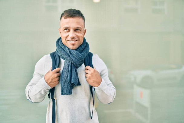 Νεαρός καυκάσιος άντρας χαμογελάει χαρούμενος φορώντας σακίδιο στην πόλη. - Φωτογραφία, εικόνα