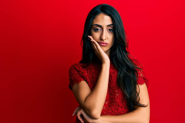 Красива іспаномовна жінка в елегантному одязі на червоному тлі думає, що виглядає втомленою і нудною з проблемами депресії з схрещеними руками
.  - Фото, зображення