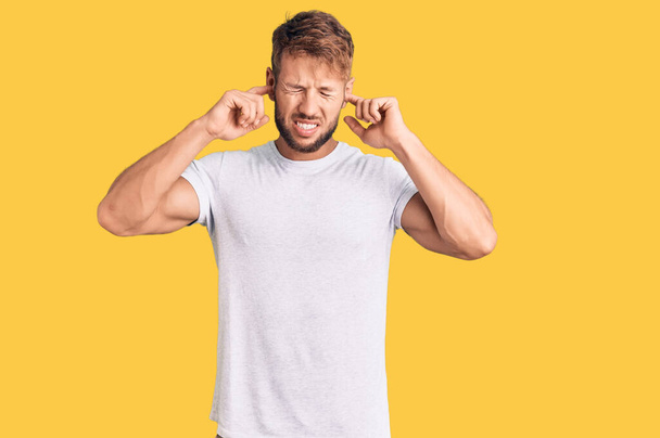 Junger kaukasischer Mann in lässigem weißem T-Shirt, der die Ohren mit den Fingern mit genervtem Gesichtsausdruck vor lauter Musik verdeckt. Gehörloses Konzept.  - Foto, Bild