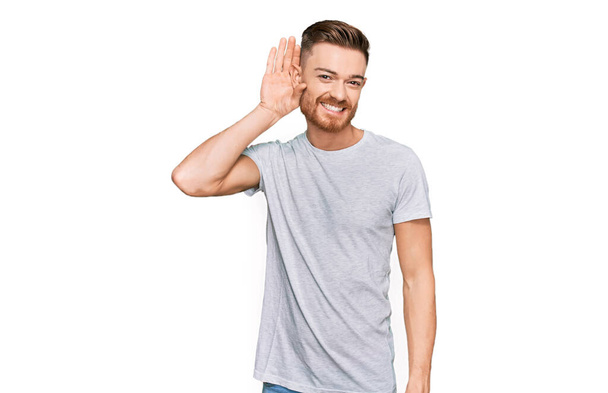 Jovem ruivo vestindo camiseta cinza casual sorrindo com a mão sobre o ouvido ouvindo um rumor ou fofoca. conceito de surdez.  - Foto, Imagem