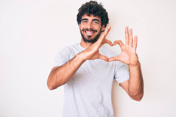 Handsome young man with curly hair and bear wearing casual tshirt smiling in love showing heart symbol and shape with hands. romantic concept.  - Photo, Image