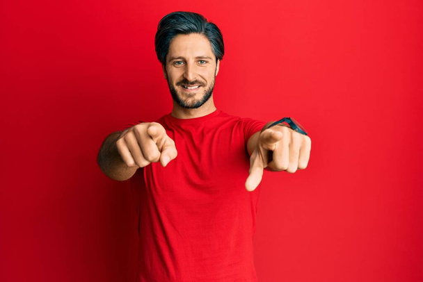 Mladý hispánec v neformálním červeném tričku ukazuje prstem na tebe a kameru, usmívá se pozitivně a vesele  - Fotografie, Obrázek