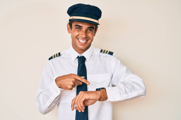 Young hispanic man wearing airplane pilot uniform in hurry pointing to watch time, impatience, upset and angry for deadline delay  - Foto, Imagem