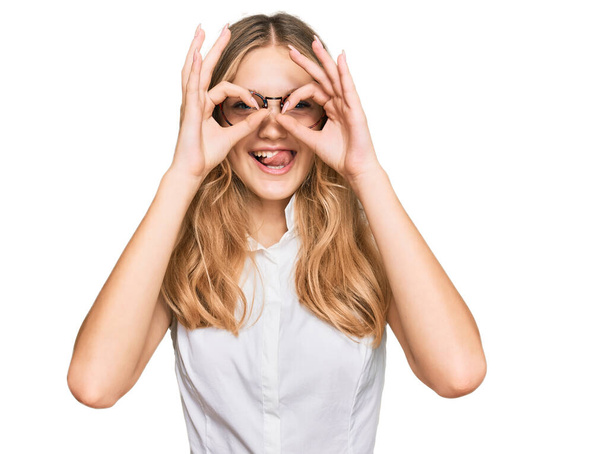 Beautiful young caucasian girl wearing casual clothes and glasses doing ok gesture like binoculars sticking tongue out, eyes looking through fingers. crazy expression.  - Photo, Image