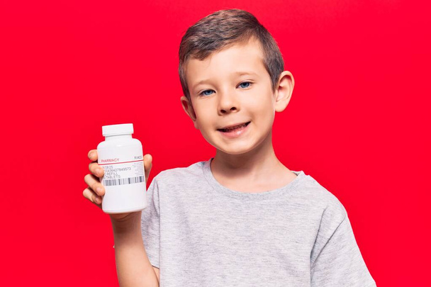 Cute blond kid holding pills looking positive and happy standing and smiling with a confident smile showing teeth  - Photo, image