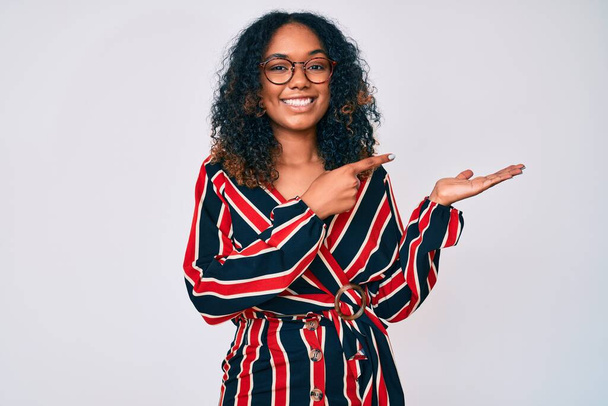Genç Afrikalı Amerikalı kadın gündelik kıyafetler ve gözlük takıyor kameraya hayret ediyor ve gülümsüyor..  - Fotoğraf, Görsel