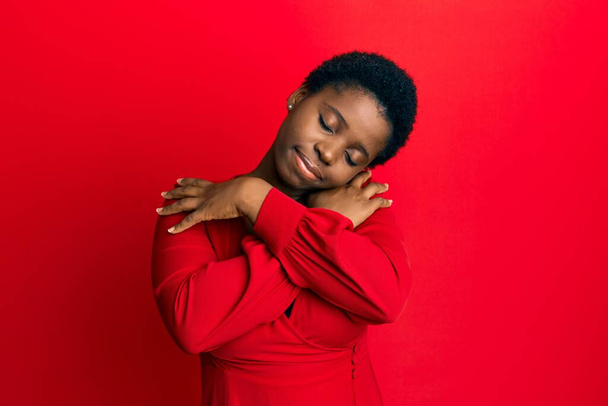 Молода африканка з афро волоссям носить повсякденний одяг на червоному тлі, обіймаючи себе щасливим і позитивним, посміхаючись впевнено. самодопомоги і самодопомоги
  - Фото, зображення