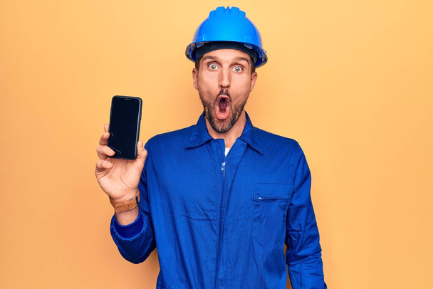 Mladý pohledný dělník v uniformě a klobouku držící smartphone ukazující obrazovku vyděšený a ohromený s otevřenými ústy pro překvapení, nevěřící tvář - Fotografie, Obrázek