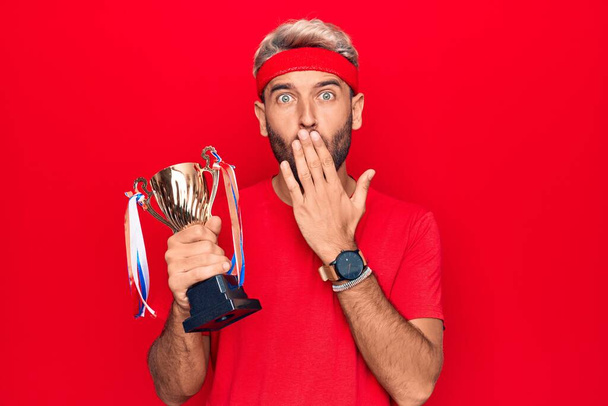 Mladý pohledný blonďatý sportovec s vousy vítězí trofej nad izolovaným červeným pozadím zakrývající ústa rukou, šokovaný a vystrašený z omylu. Překvapený výraz - Fotografie, Obrázek