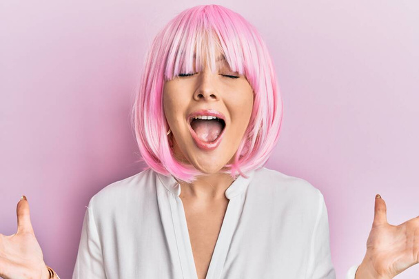 Junge blonde Frau mit pinkfarbener Perücke feiert verrückt und erfolgsverrückt mit erhobenen Armen und geschlossenen Augen, die aufgeregt schreien. Siegerkonzept  - Foto, Bild