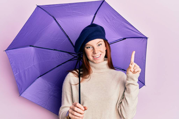 Joven chica de cabeza roja sosteniendo paraguas púrpura con boina fresca sorprendida con una idea o pregunta apuntando con el dedo con la cara feliz, número uno  - Foto, imagen