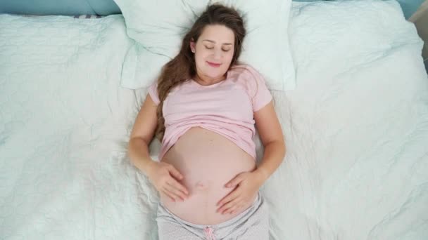 Όμορφη έγκυος γυναίκα με πιτζάμες να ξεκουράζεται στο κρεβάτι και να χαϊδεύει την κοιλιά της. Έννοια της εγκυμοσύνης, προετοιμασία και αναμένουν παιδί στο σπίτι - Πλάνα, βίντεο