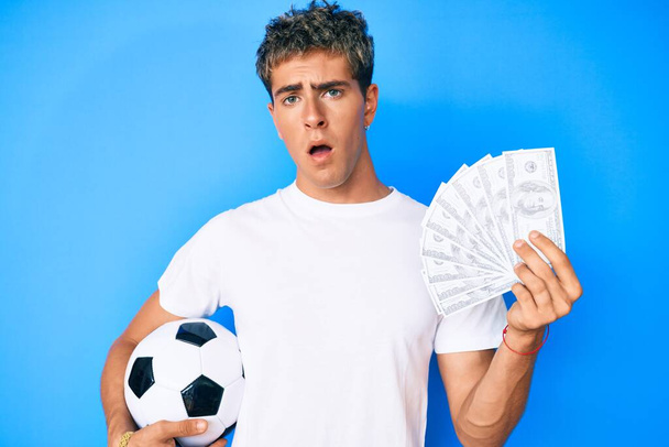 Elinde futbol topu ve dolar banknotları olan genç yakışıklı adam, şaşkın, şüpheci ve alaycı görünüyor, ağzı açık kalınca şaşırıyor.  - Fotoğraf, Görsel