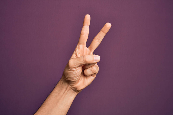 Mano de joven caucásico mostrando los dedos sobre fondo púrpura aislado contando número 2 mostrando dos dedos, gestos de victoria y símbolo ganador - Foto, imagen