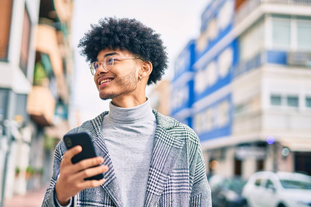 Joven hombre de negocios afroamericano sonriendo feliz usando el teléfono inteligente en la ciudad. - Foto, imagen
