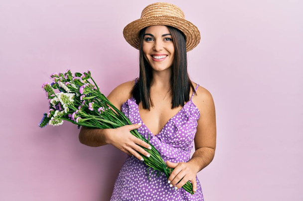 Young brunette woman wearing summer dress holding bouquet of flowers looking positive and happy standing and smiling with a confident smile showing teeth  - Photo, Image