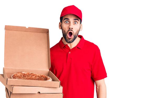 Junger gutaussehender Mann mit Bart trägt Lieferuniform, hält Schachteln mit Pizza in der Hand und staunt mit offenem Mund über Überraschung, ungläubiges Gesicht  - Foto, Bild