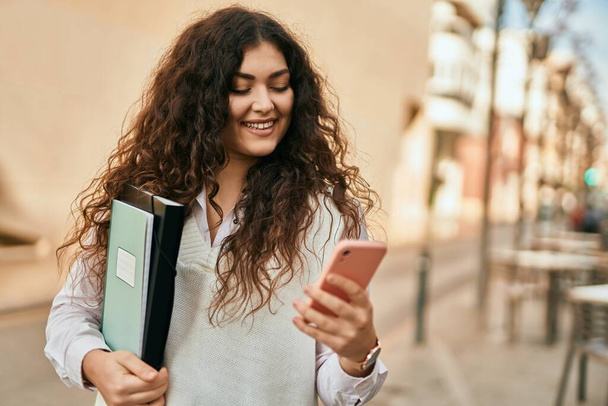 Νεαρή ισπανόφωνη γυναίκα χαμογελά ευτυχισμένη χρησιμοποιώντας smartphone στην πόλη. - Φωτογραφία, εικόνα