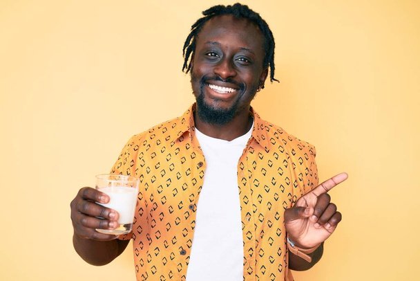 Jovem afro-americano com tranças segurando vidro de leite sorrindo feliz apontando com a mão e o dedo para o lado  - Foto, Imagem