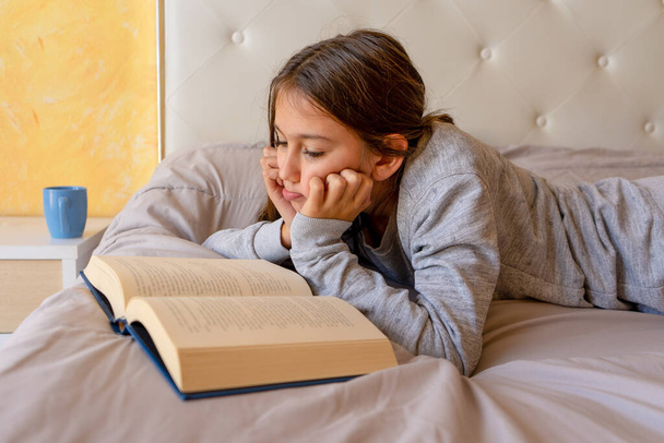 Słodka dziewczynka czytająca książkę leżącą na łóżku w jej pokoju. Wybiórcze skupienie. Koncepcja uczenia się - Zdjęcie, obraz