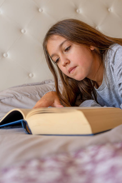 Cute little girl reading a book lying on the bed in her room. Selective focus. Learning concept - Zdjęcie, obraz