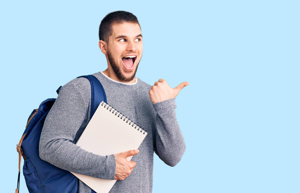 Joven hombre guapo con mochila de estudiante sosteniendo portátil apuntando el pulgar hacia el lado sonriendo feliz con la boca abierta  - Foto, Imagen