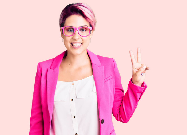 Fiatal gyönyörű nő rózsaszín haj visel üzleti kabát és szemüveg mosolyog boldog arc kacsintott a kamera csinál győzelem jele. Második..  - Fotó, kép